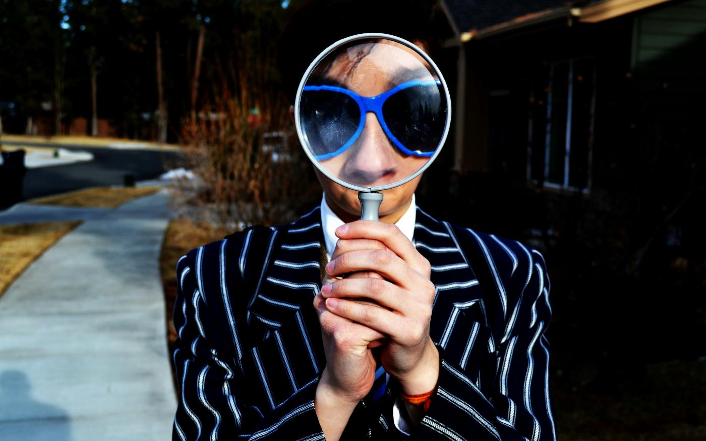 person using magnifying glass enlarging the appearance of his nose and sunglasses by Marten Newhall courtesy of Unsplash.